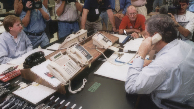 This colorful photo shows reporters filming Hurricane Specialist Richard Pasch making a hurricane conference call with the National Weather Service New Orleans/Baton Rouge Office during Hurricane Katrina in 2005. On the right, a man sits on one of a bank of phones with his back to us. Reporters with cameras and microphones crowd around the table.
